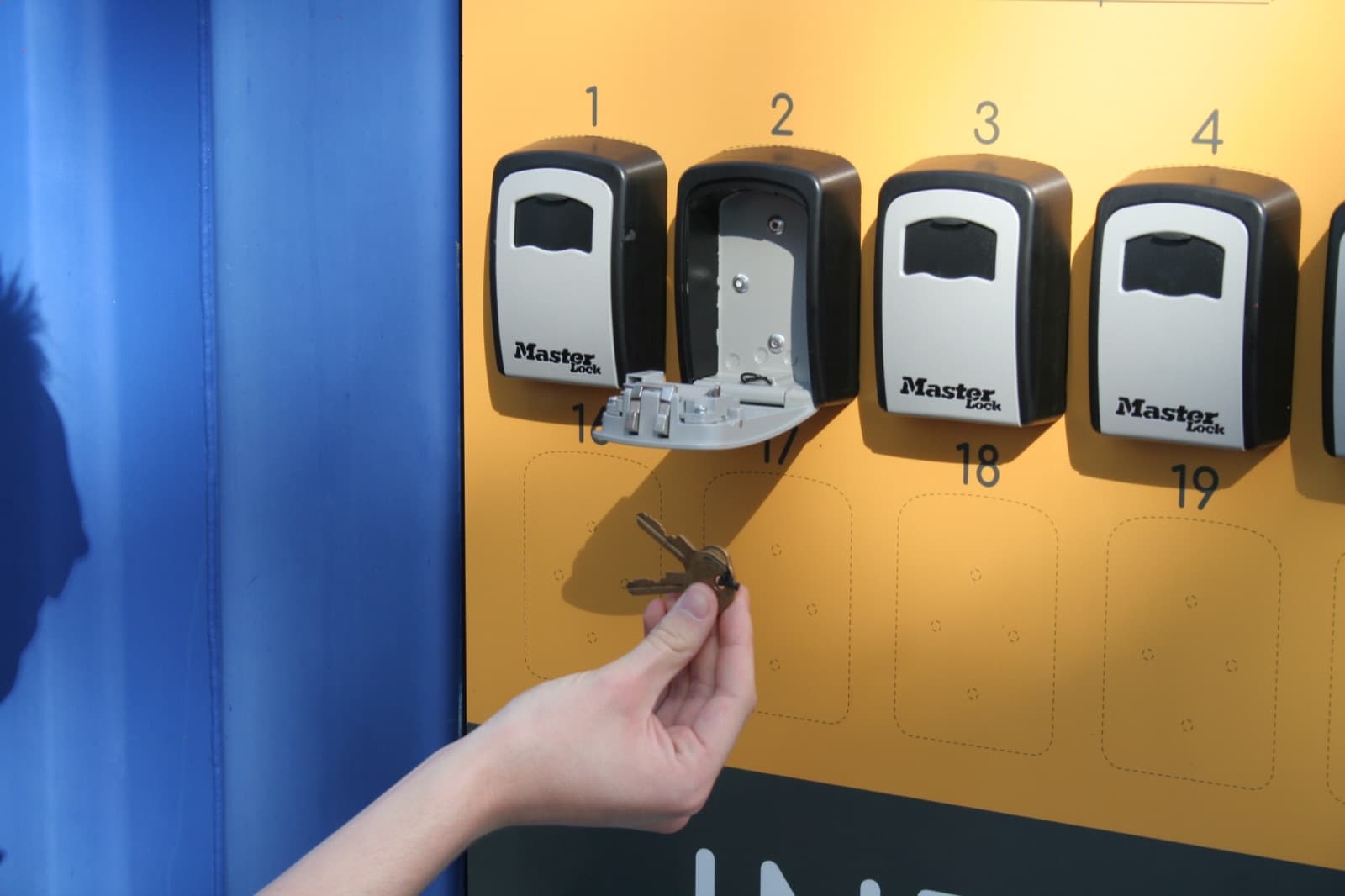 Quatre boîtes à clés à code numérotées, celle du box n°2 ouverte, une main tenant la clé du cadenas en dessous